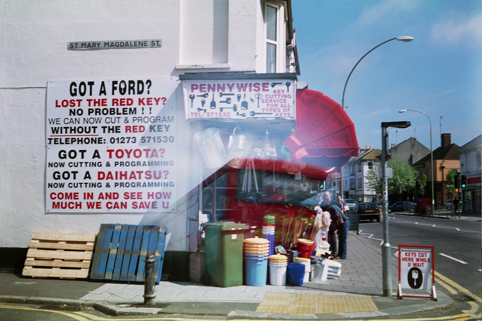 a corner shop displaying lots of things like watering cans, plastic buckets, etc with a sign saying they can offer key cutting services for ford. The shop's name is Pennywise. There are also some pallets leaning against the wall. There is a road sign saying 'St Magdalene Street'. There is blue sky and we see the main road going further upwards towards the distance.